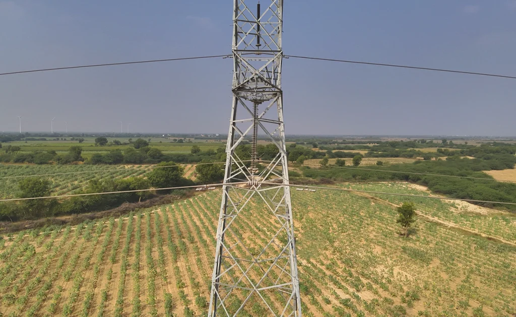 Aerial drone photograph of a 220kV lattice transmission tower rising above agricultural farmland in India, showing insulator strings, bundled conductors, and clear right-of-way corridor for drone-based inspection and LiDAR survey.