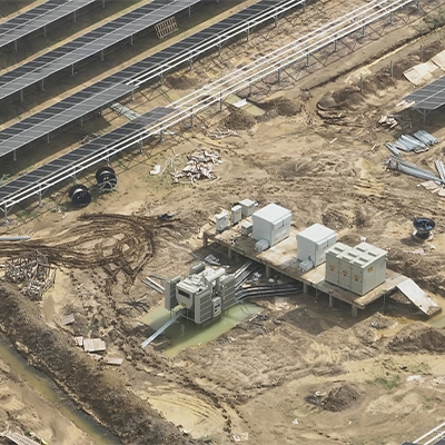 Aerial drone view of a solar inverter station zone mid-construction — showing cable trenching, waterlogged civil works, installed transformer units, and adjacent completed module rows, all visible in a single flight.