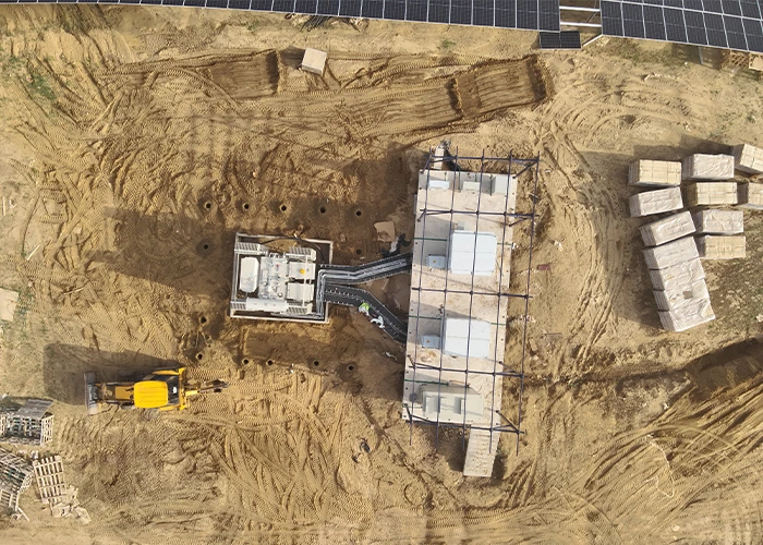 Overhead drone view of solar inverter station construction with a yellow excavator, precast concrete blocks, steel framework, and earthwork — documenting construction-phase progress at equipment level.