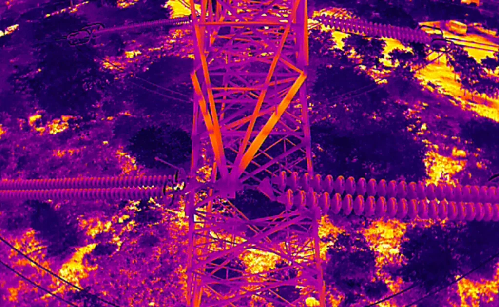 Thermal infrared drone image of a high-voltage transmission tower showing radiometric heat mapping of insulators, conductor clamps, and lattice structure for predictive fault detection in India.