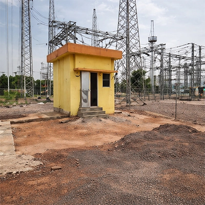 Ground-level photograph of a yellow painted control room cabin at a solar farm grid substation in India, with high-voltage transmission towers, switchyard gantry structures, and red laterite soil earthworks visible in the background.
