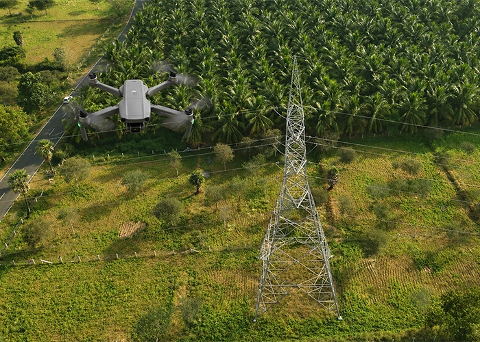 Drone inspecting high voltage transmission tower in India surrounded by agricultural land and coconut plantation