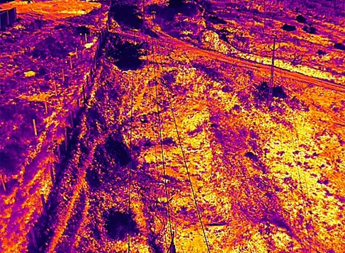 Aerial thermal image of overhead cables or wires crossing terrain with embankments in purple-to-yellow heat palette