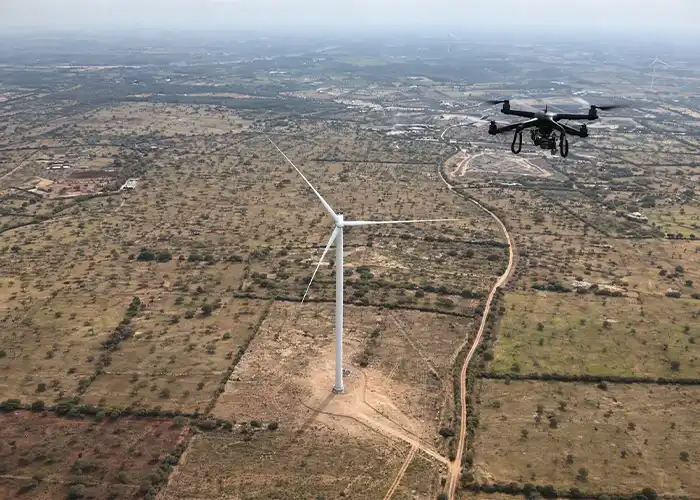 Drone conducting aerial survey alongside wind turbine over dry agricultural landscape in India, additional turbines on horizon