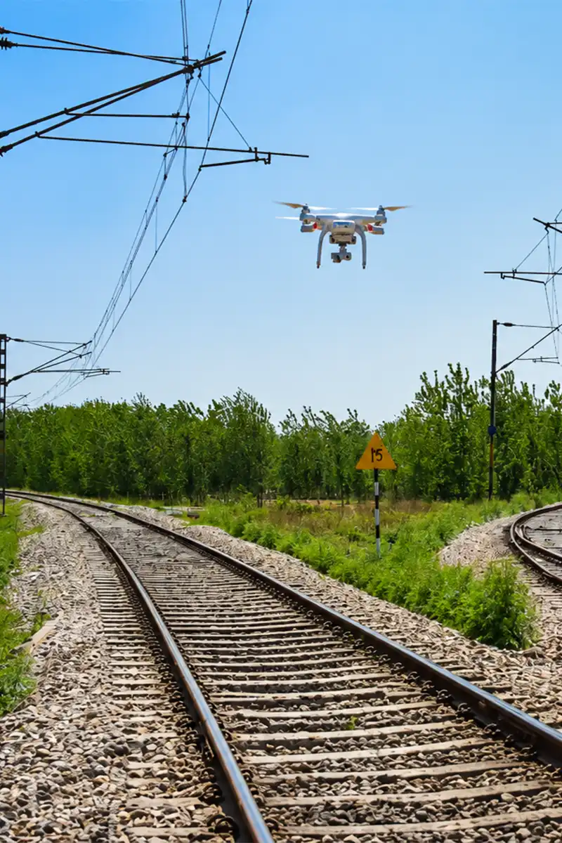 White quadcopter drone flying over electrified railway tracks with overhead catenary equipment and speed restriction sign