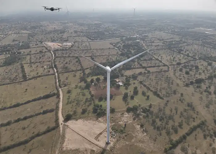 Small drone in flight above white wind turbine during aerial inspection over agricultural landscape in India