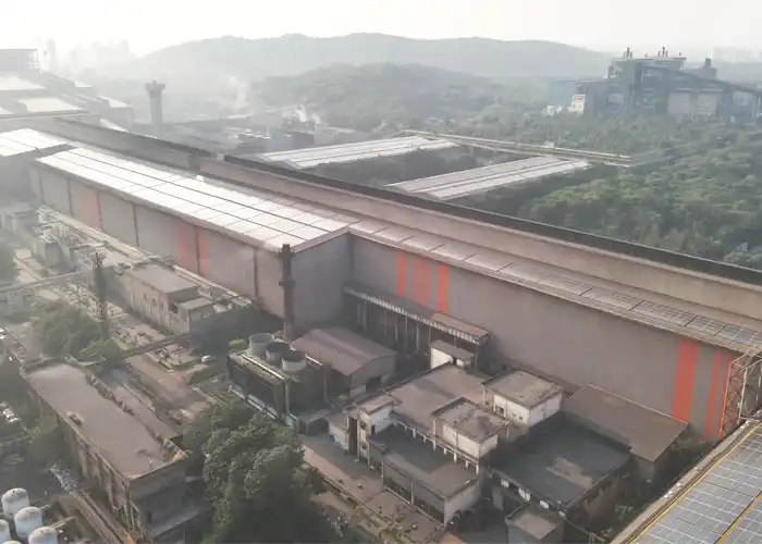 Aerial drone view of large heavy industrial plant with solar panels on rooftop, forested hills visible in hazy background