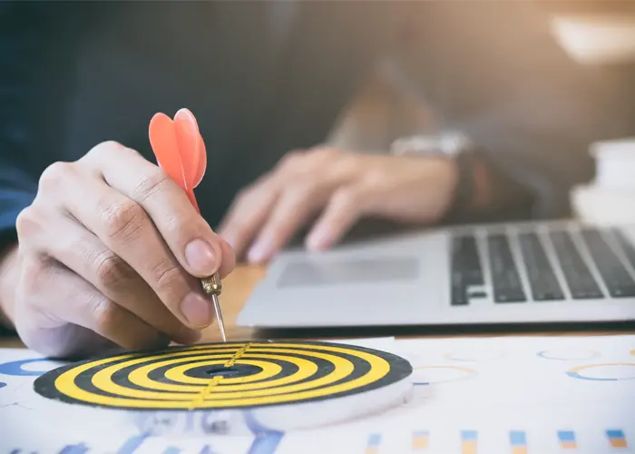 Hand placing dart on bullseye target at desk with charts and laptop precision accuracy concept