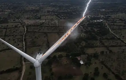 Wind turbine blade struck by lightning, bright bolt visible along blade length against dark stormy sky over agricultural landscape