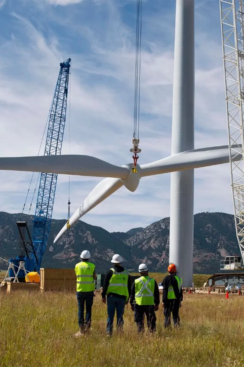 Four engineers observing wind turbine rotor assembly lifted by blue lattice crane during installation with mountain backdrop